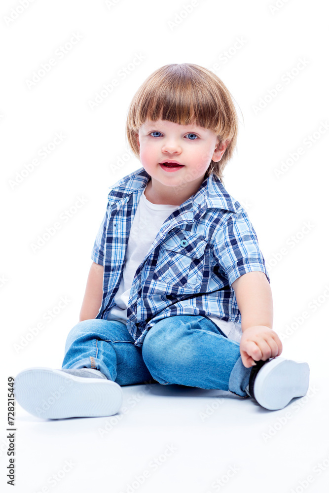 Smiling little boy 2 years old sitting on the floor. Child in a plaid shirt and jeans. Happy childhood. White background. Vertical. Smiling little boy 2 years old sitting on the floor. Child in a plaid shirt and jeans. Happy childhood. White background. Vertical.