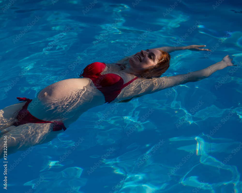 Top view of pregnant woman floating in pool in red bikini. 