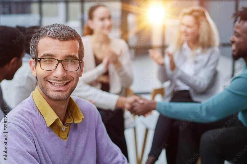Obraz premium A happy young businessman, in a purple sweater, sits against the background of his colleagues
