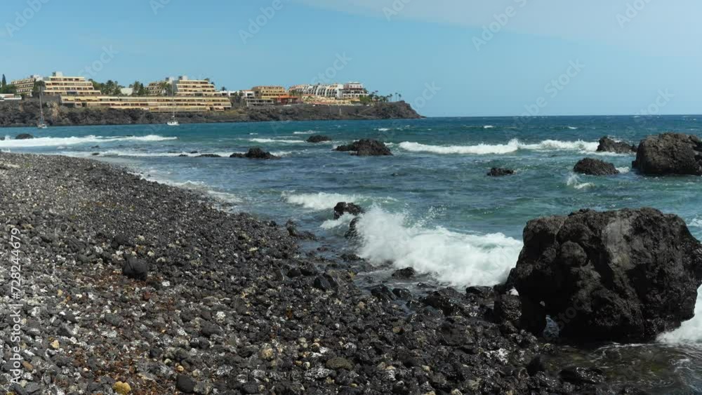 Establishing view of ocean waves crashing across volcanic black rock pebble beach with hotels in distance Establishing view of ocean waves crashing across volcanic black rock pebble beach with hotels in distance
