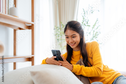 Happy young Asian woman sitting on sofa holding mobile phone using cellphone technology doing ecommerce shopping  buying online  texting messages relaxing on couch in cozy living room at home
