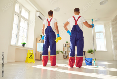 Backside full length portrait of a two men in uniform of a cleaning company. Equipped with mop and various equipment for housekeeping, this image captures cleaning service staff engaged in cleanup.