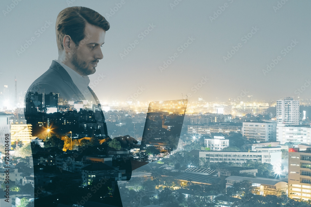 Attractive young european businessman using laptop while standing on night city background. Success, technology, communication, future and tomorrow concept. Double exposure. - obrazy, fototapety, plakaty 