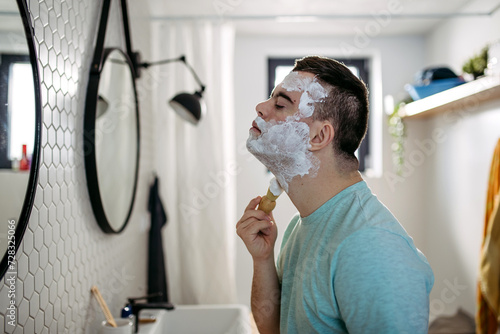 Young man with down syndrome learning how to shave, applying shaving foam all over his face, looking at mirror. photo