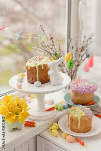 Cakes decorated with flowers and eggs near Easter bunny toy on window sill