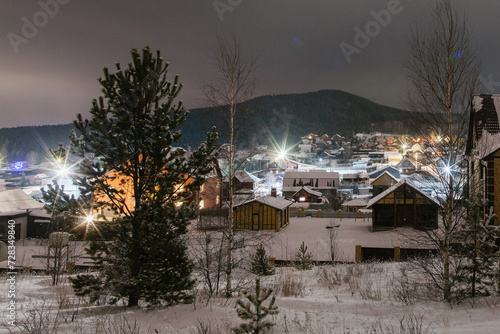 Mountain landscape with village in winter, houses and pine trees covered snow at night. Scenery of ski resort with lights. Theme of travel.