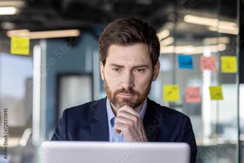 Serious concentrated thinking financier businessman at workplace preparing electronic financial report, mature experienced man in business suit typing laptop keyboard, reading report, inside office.