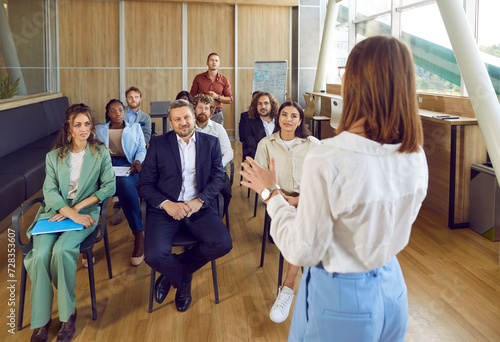 Confident female business coach teaches small group of people new business knowledge. People are sitting on chairs in office of business center listening to woman standing with her back to camera. photo