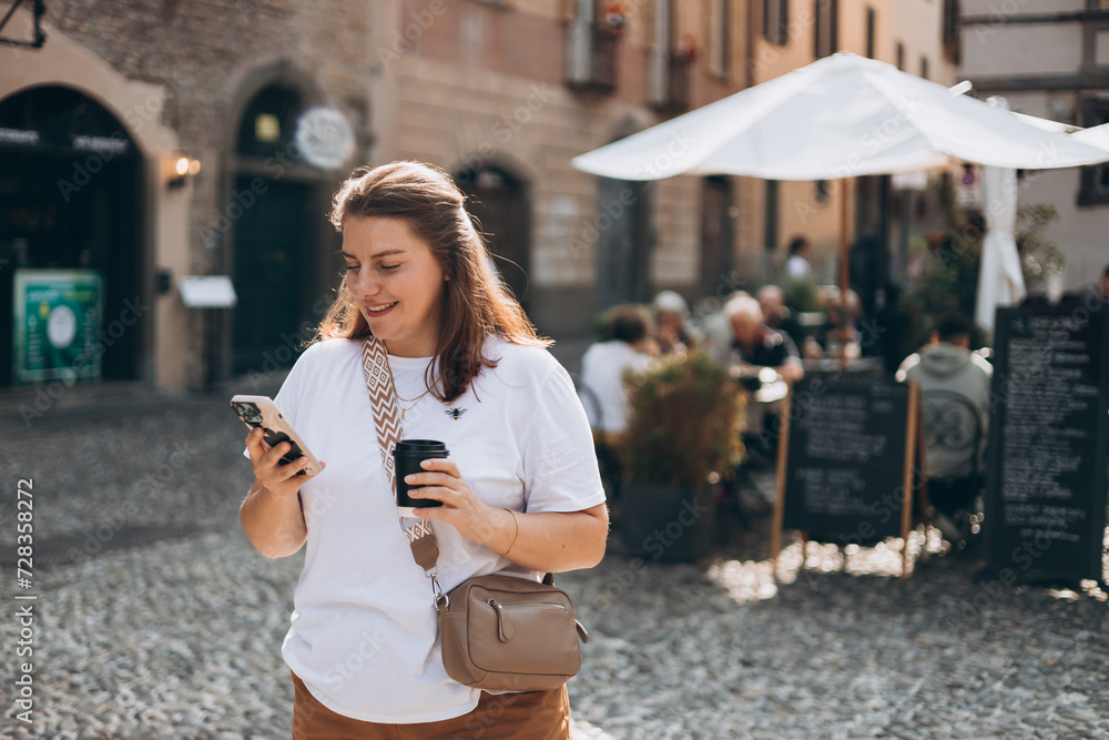 Happy young Caucasian woman in good mood walks with coffee and phone around city during day. Lifestyle concept. 30s brunet girl holding takeaway coffee cup near cafe in old town