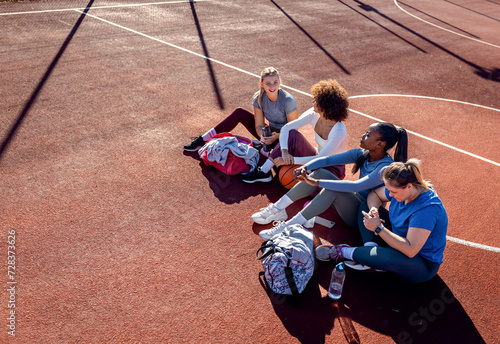 Diverse group of young woman sitting on court resting afrer playing basketball outdoors. photo