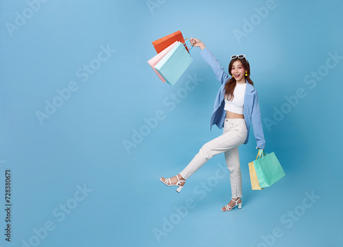 Full length young asian shopper woman walking and holding shopping bag feeling happy excited for discount promotion isolated on blue copy space background.