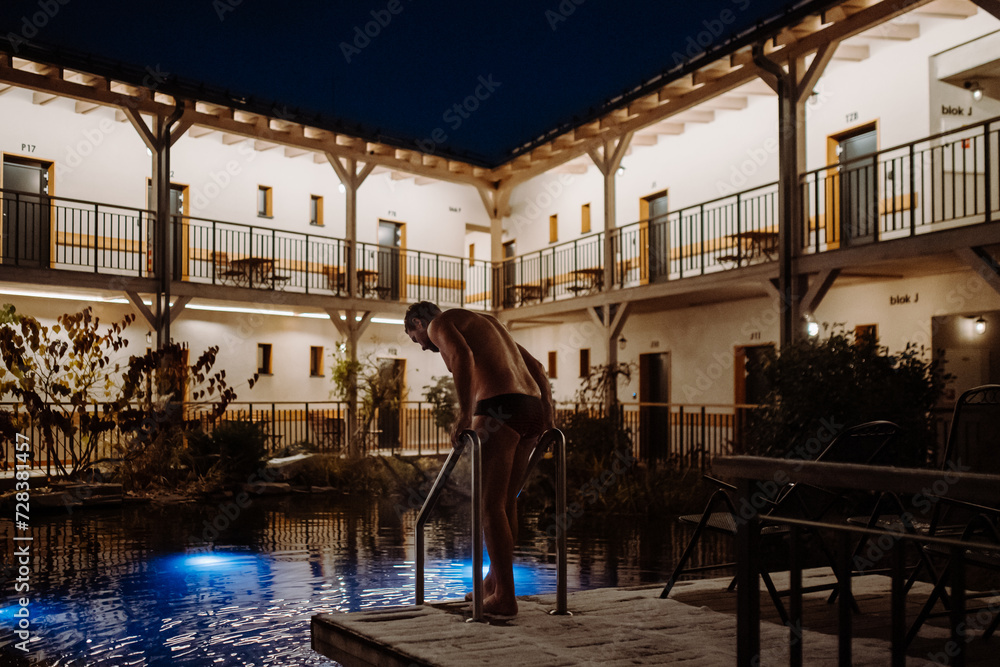Mature man swimming in outdoor pool at night, enjoying calmness and empty pool. Wellness weekend in hotel.