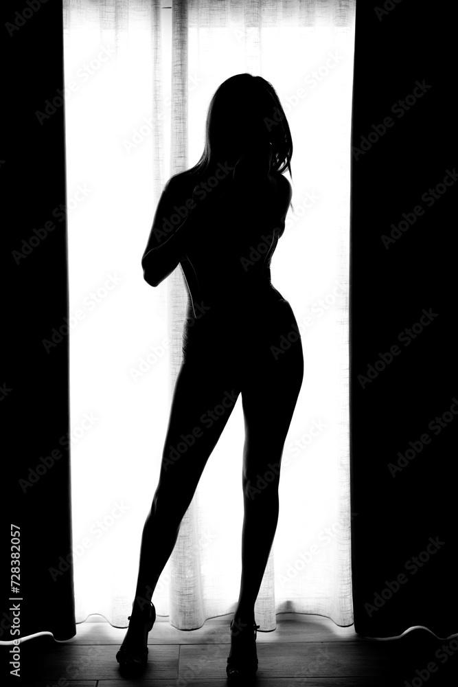 Silhouette of an attractive woman with wide hips in a window with curtains on her sides.
