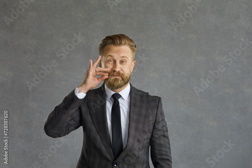 Confident caucasian businessman pretends to lock his mouth promising to keep a secret. Middle aged man shows a gesture of silence on a gray concrete background. Concept of secrecy and silence.
