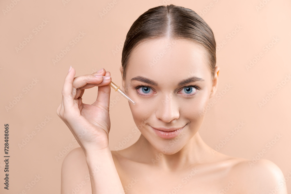 Beautiful young woman applying serum onto her face on beige background in studio