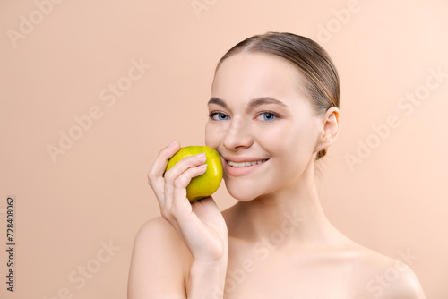 Young woman holding green apple, portrait with bare shoulders on beige background