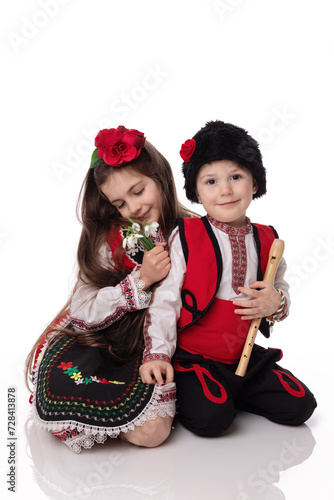 Bulgarian kids boy and girl in traditional ethnic folklore clothes, spring flowers snowdrops, flute and red white wool bracelet martenitsa symbol March holiday Baba Marta, Bulgaria