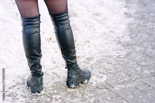 Woman legs with knee-high boots, winter outfit. Woman legs in high boots and black pantyhose stands on snowy sidewalk, fashion footwear