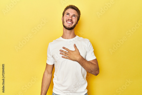 Young Caucasian man on a yellow studio background laughs out loudly keeping hand on chest.