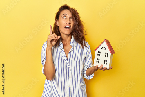 Middle aged woman holding a miniature house on yellow backdrop pointing upside with opened mouth.