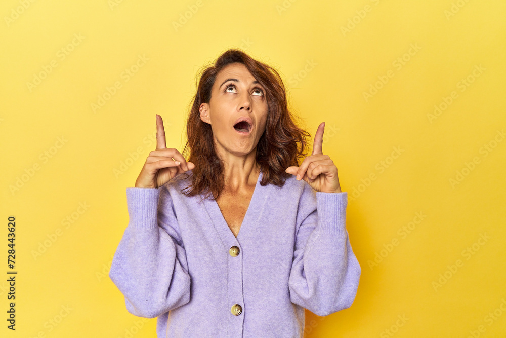 Middle-aged woman on a yellow backdrop pointing upside with opened mouth. Middle-aged woman on a yellow backdrop pointing upside with opened mouth.