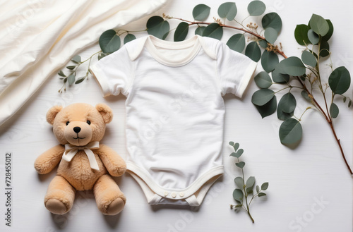 White cotton baby short sleeve bodysuit with cute teddy bear, eucalyptus branch on white ivory blanket throw background.