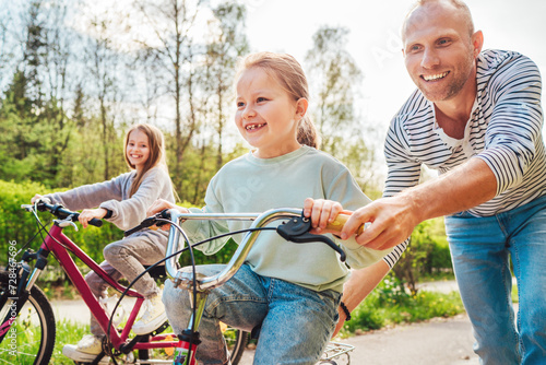 Smiling father with two daughters during outdoor walk. He teaching younger girl to ride a bicycle. They enjoy togetherness in the summer city park. Happy childhood concept image..
