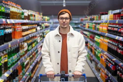 Male shopper with glasses and beanie holding a cart in a drink aisle. Portrait of a man with a shopping cart in a supermarket looking at the camera against the background of shelves with carbonated photo