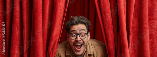 A joyful man with glasses and curly hair expressing surprise and excitement behind red stage drapes. photo
