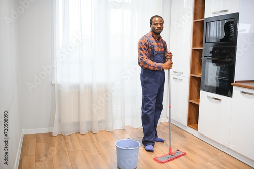 Professional cleaner in blue uniform washing floor and wiping dust from the furniture in the living room of the apartment. Cleaning service concept