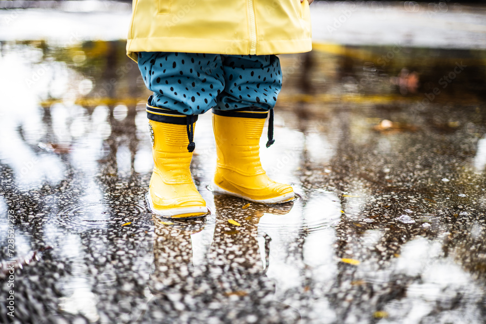Small infant boy wearing yellow rubber boots and yellow waterproof raincoat standing in puddle on a overcast rainy day. Child in the rain Small infant boy wearing yellow rubber boots and yellow waterproof raincoat standing in puddle on a overcast rainy day. Child in the rain