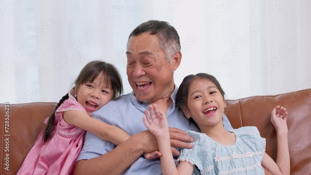 Happy Asian senior grandfather sits on a couch with his granddaughter and plays with his granddaughter in the living room at home, The Concept of family having fun in their house Happy Asian senior grandfather sits on a couch with his granddaughter and plays with his granddaughter in the living room at home, The Concept of family having fun in their house