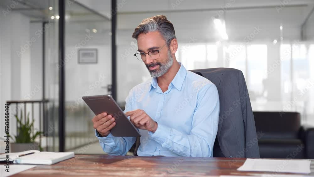 Middle aged Hispanic business manager ceo using tablet pc computer app. Smiling Latin or Indian mature man businessman holding gadget device sit at table in office working online in internet indoors. Middle aged Hispanic business manager ceo using tablet pc computer app. Smiling Latin or Indian mature man businessman holding gadget device sit at table in office working online in internet indoors.