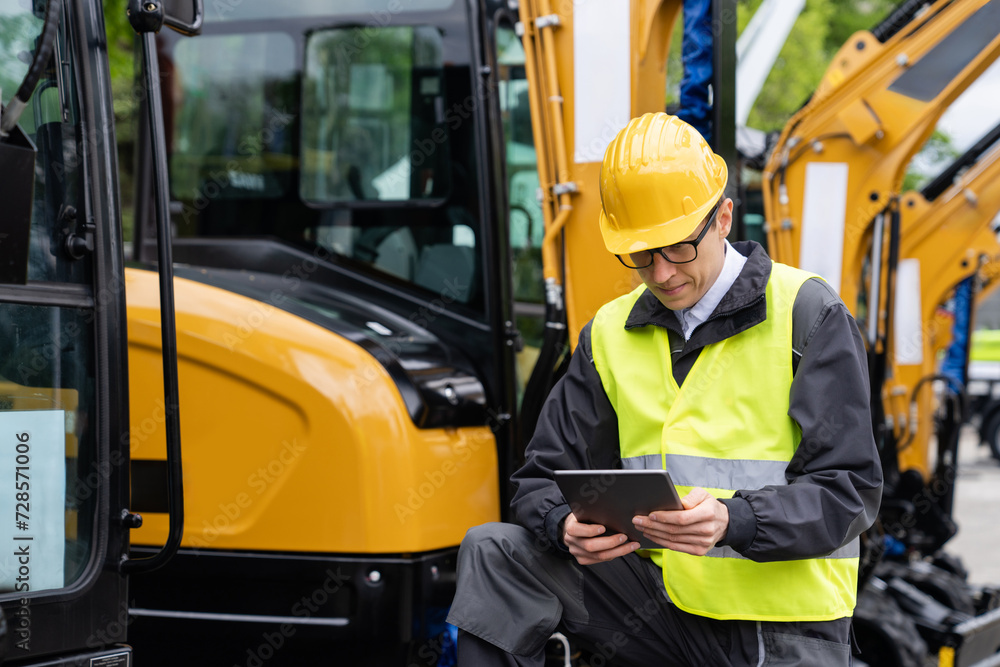 Engineer in a helmet with a digital tablet stands next to construction excavators.. Engineer in a helmet with a digital tablet stands next to construction excavators..