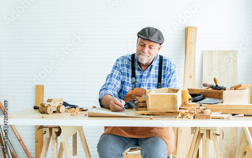 Senior Caucasian happy retired old male woodworker or carpenter smiling, wearing check shirt with apron, vintage hat, creating DIY wooden mini figure toy for decoration as hobby after retirement.