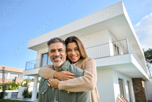 Happy smiling mature older family couple new property buyers modern dream luxury villa owners standing outside house, affectionate senior middle aged man and woman in love hugging outdoor. Portrait.