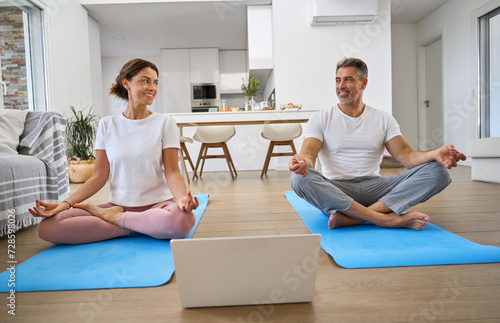 Happy mature couple learning online yoga class on computer at home. Smiling senior serene healthy middle aged man and woman doing training exercises meditating together using laptop in living room. photo