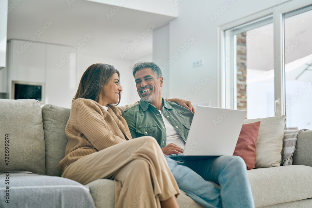 © insta_photos - Happy middle aged couple using laptop computer relaxing on couch at home. Smiling mature man and woman talking having fun laughing with device sitting on sofa in sunny living room. Candid shot.