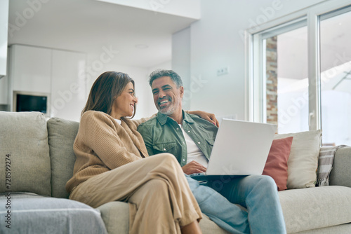Happy middle aged couple using laptop computer relaxing on couch at home. Smiling mature man and woman talking having fun laughing with device sitting on sofa in sunny living room. Candid shot.