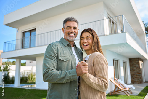 Portrait of happy smiling mature older family couple new property buyers modern dream luxury villa owners standing outside house, affectionate senior middle aged man and woman in love hugging outdoor.
