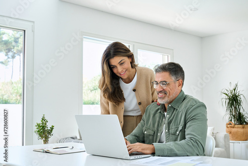 Smiling middle aged senior man working on computer sitting at table with wife standing nearby in living room. Happy mature older couple using laptop technology at home. Authentic candid shot.