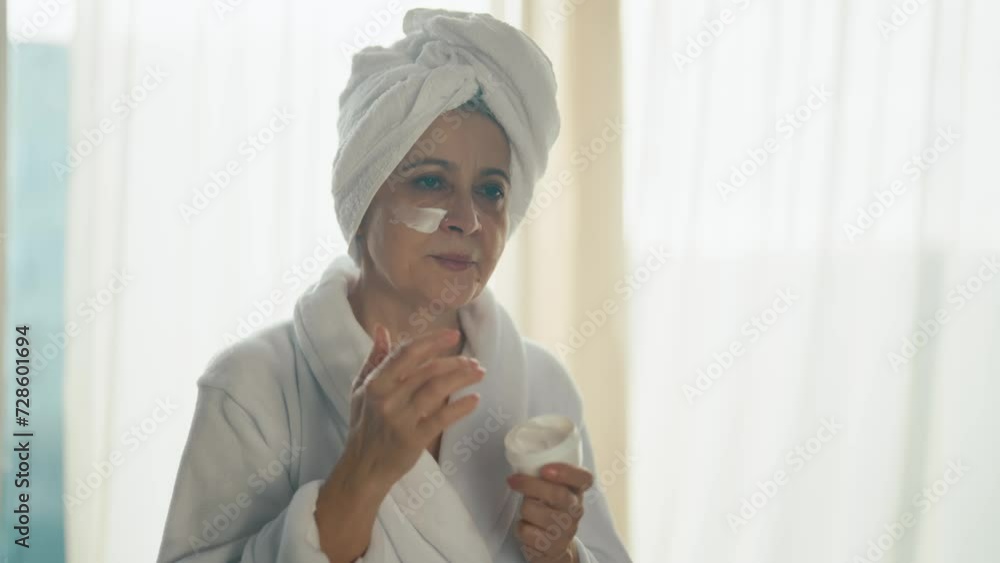 Contented senior gray haired woman applying clay face mask for moisturizing skin. Smiling old lady in white bathrobe. Skin care, home lifting spa cosmetology, domestic procedures treatment concept.