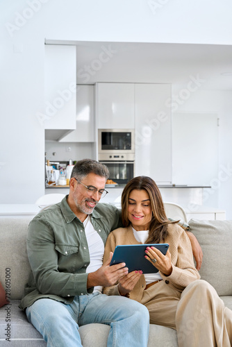 Vertical shot of relaxed happy middle aged couple mature man and woman hugging sitting on sofa at home relaxing on couch using digital tab buying online browsing internet in modern house living room.