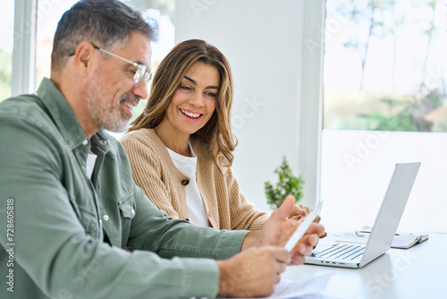 Happy middle aged mature couple using laptop counting taxes refund receipts to save money at home. Old man and woman paying bills online planning financial budget savings calculating payment at table. photo