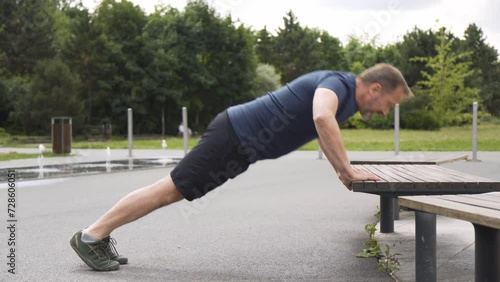 A handsome middle-aged Caucasian man does incline push-ups against a bench in a park photo