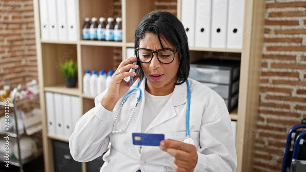 A concerned hispanic woman doctor examines a credit card while on a phone call in a clinic office. A concerned hispanic woman doctor examines a credit card while on a phone call in a clinic office.
