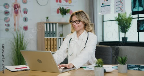 Portrait of woman doctor in glasses and hospital uniform which typing on computer in medical workroom