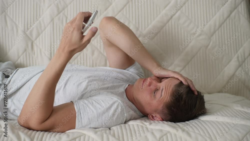 Happy tired sleepy young man yawning while lying on sofa watching video online on smartphone in living room at home. Exhausted male finishing texting on phone, gives yawny sigh and sinks into sleep. Happy tired sleepy young man yawning while lying on sofa watching video online on smartphone in living room at home. Exhausted male finishing texting on phone, gives yawny sigh and sinks into sleep.