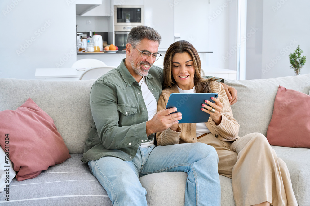 Happy middle aged couple using digital tablet relaxing on couch at home. Smiling mature man and woman holding tab computer browsing internet on pad technology device sitting on sofa in living room. Happy middle aged couple using digital tablet relaxing on couch at home. Smiling mature man and woman holding tab computer browsing internet on pad technology device sitting on sofa in living room.