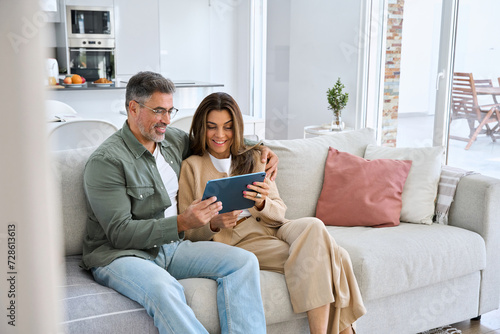 Happy middle aged couple using digital tablet relaxing on couch at home. Smiling mature man and woman holding tab browsing internet on pad device sitting on sofa in living room. Authentic candid photo photo